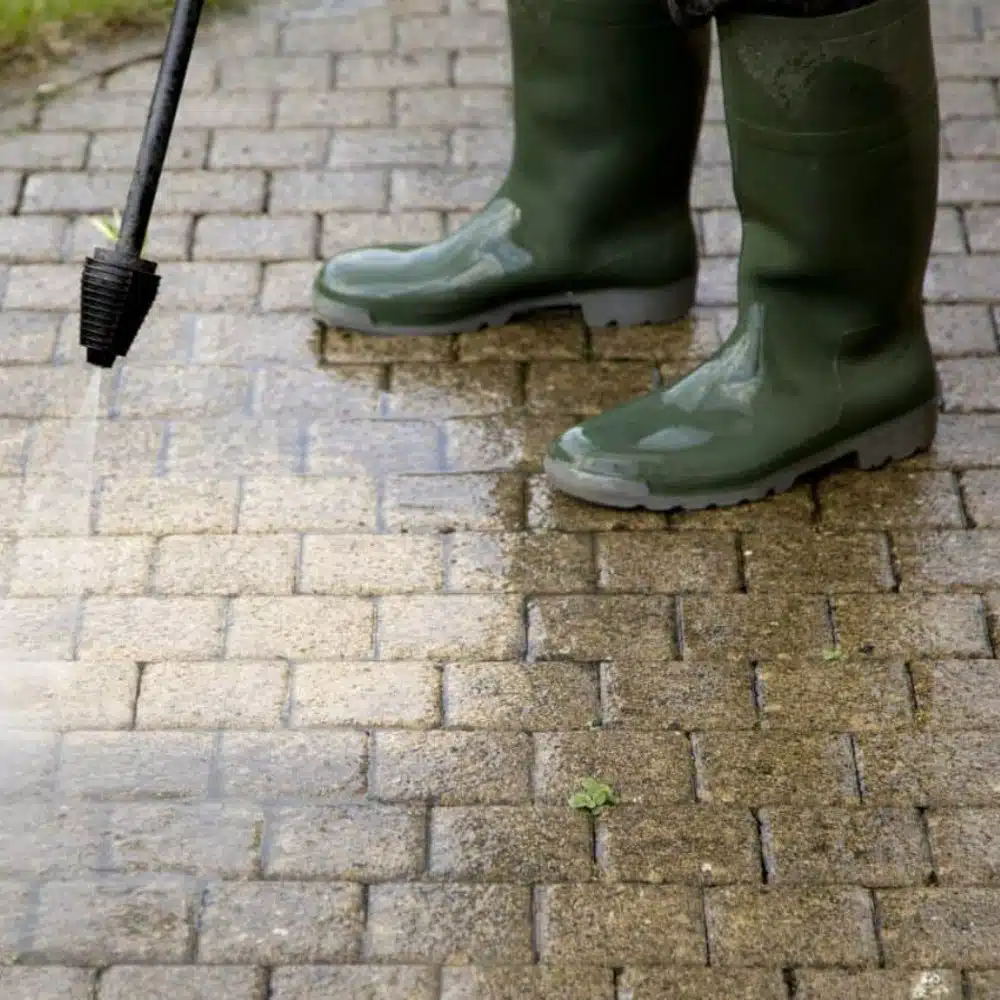 Power Washing paver Green boots stand on a brick paver patio, half of which has been pressure washed clean. The other half shows dirt and moss. Illustrates the results of paver sealing and cleaning.