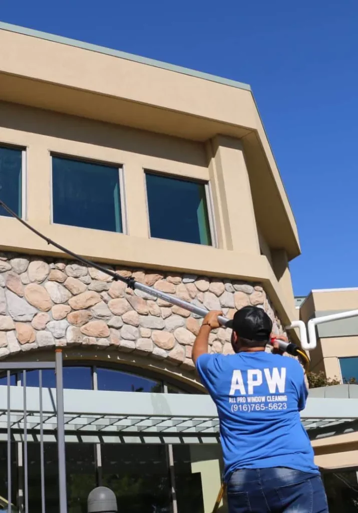 Top Rated Window Cleaning Roseville CA Mob A window cleaner uses a long pole to clean high windows on a building with stone accents. The blue sky provides a backdrop for this home window cleaning service.